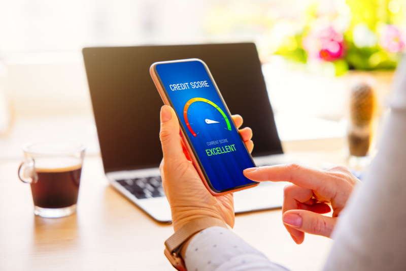 A person holds a smartphone displaying a credit score gauge with the status marked as Excellent. A laptop, a cup, and blurred plants are visible in the bright background.