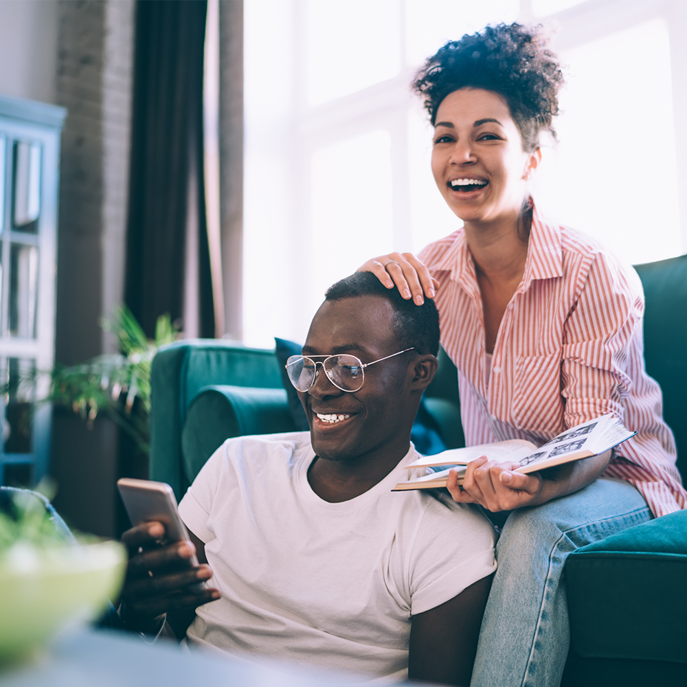 A smiling couple relaxes in a bright living room. The man sits on the floor looking at his phone, while the woman, holding a magazine, sits on the couch behind him, resting her hand on his shoulder.